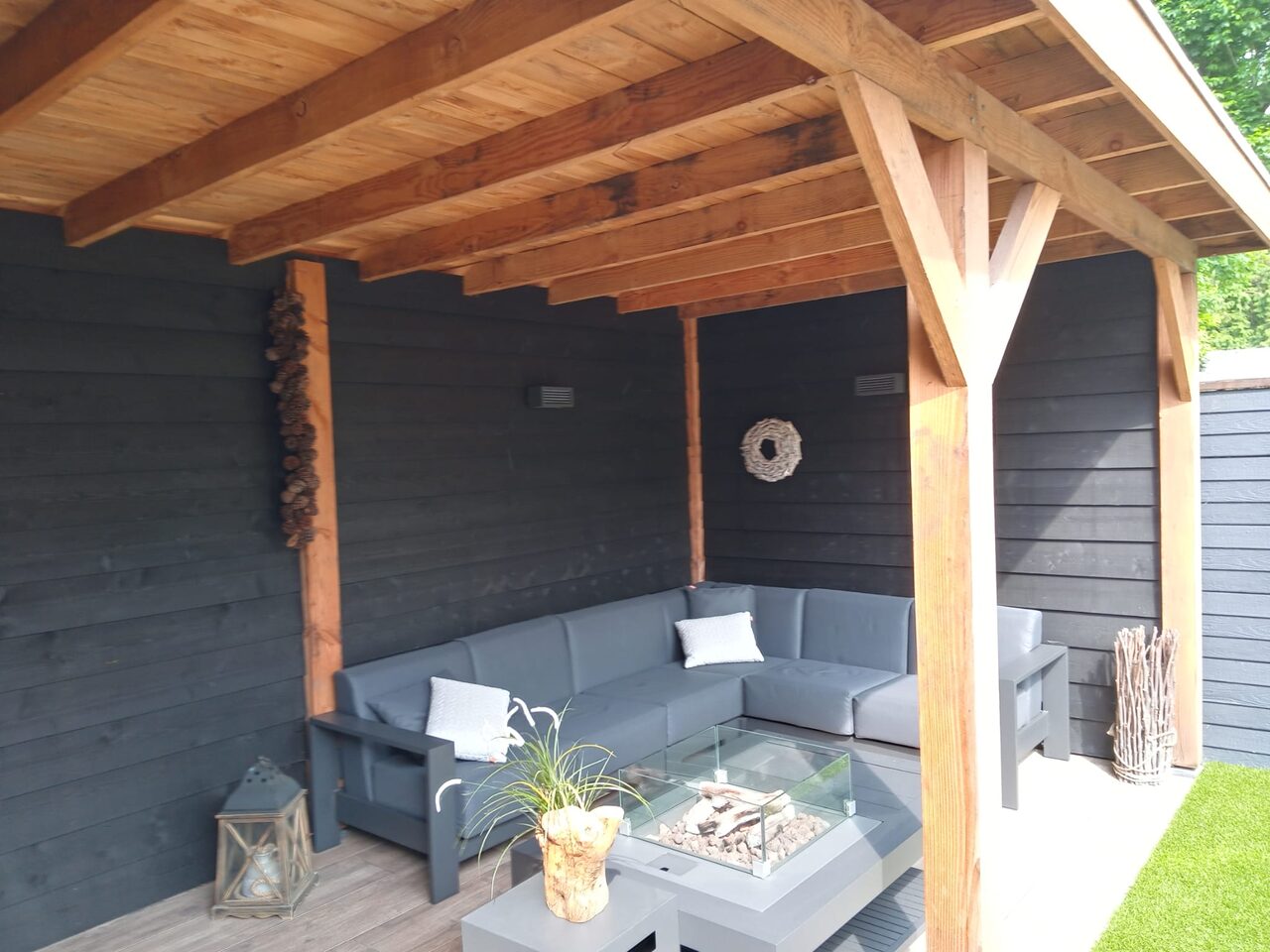 Loungeset met glazen vuurtafel onder een houten overkapping met zwarte wanden