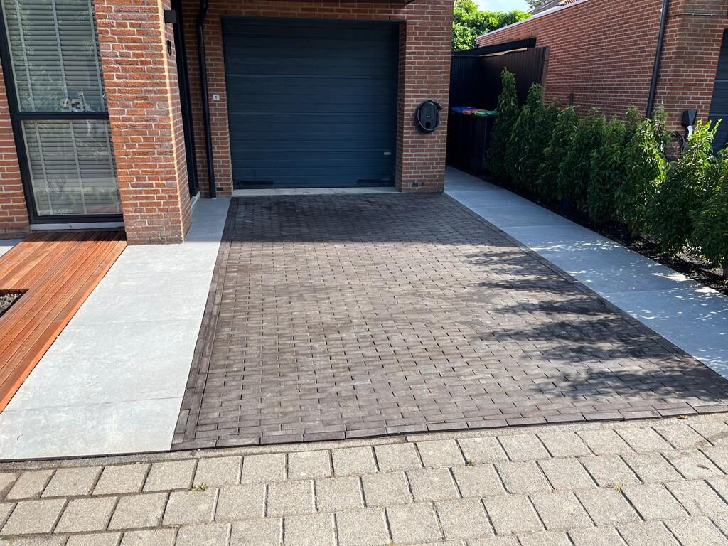 Oprit met klinkerbestrating naar de garagedeur, lichte tegelstrook en hardhouten vlonder