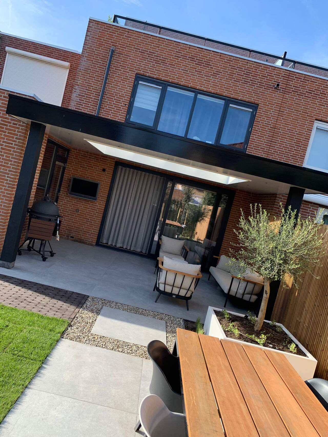 Achtertuin met overkapping, loungeset, houten eettafel en olijfboom in witte bak