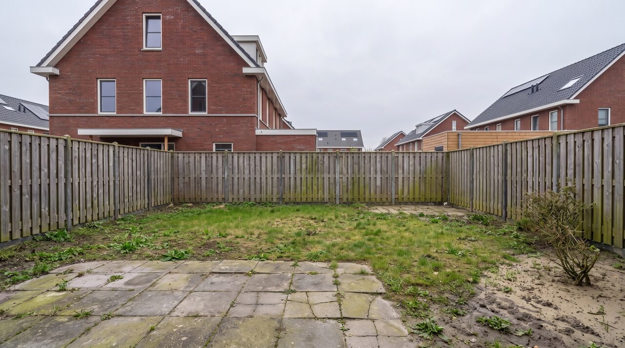 Karige tuin voor de aanleg: kaal gras, oude tegels en een vergrijsde schutting