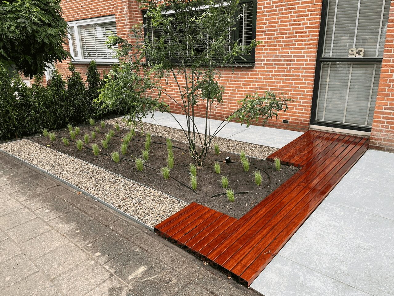 Voortuin met hardhouten vlonderpad, lichte tegels, grindstrook en een pas aangeplante border met siergrassen en meerstammige boom